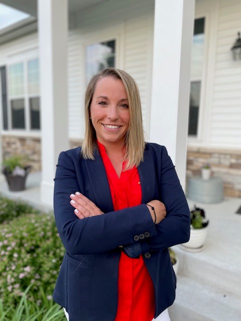 emily karadak realtor standing with arms crossed in front of a white or beige home